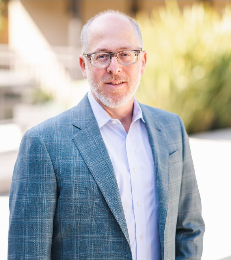 A man with glasses, a beard, and a plaid jacket stands outdoors, facing the camera.